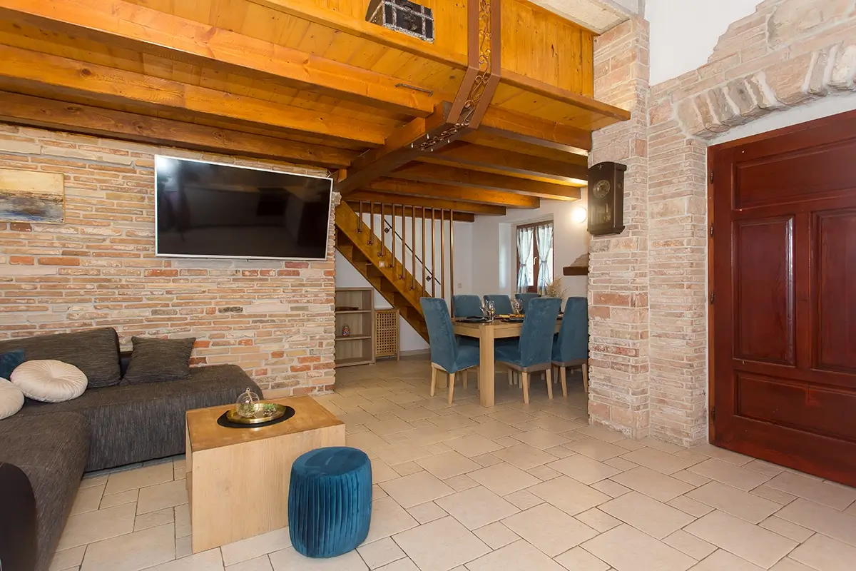 Living room with a large sofa, television, wooden stairs to the second floor, dining table, and brown wooden entrance doors.